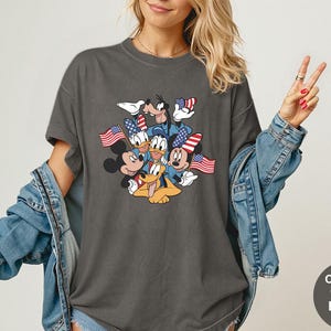 May include: Dark gray t-shirt featuring a graphic of Mickey Mouse, Minnie Mouse, Donald Duck, Goofy, and Pluto, each holding or wearing American flags. The shirt is a Comfort Colors Pepper tee.