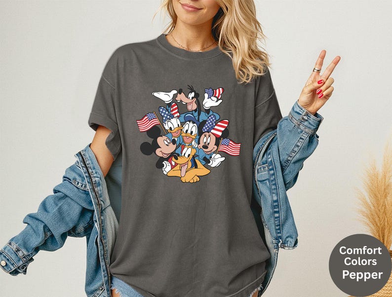 May include: Dark gray t-shirt featuring a graphic of Mickey Mouse, Minnie Mouse, Donald Duck, Goofy, and Pluto, each holding or wearing American flags. The shirt is a Comfort Colors Pepper tee.