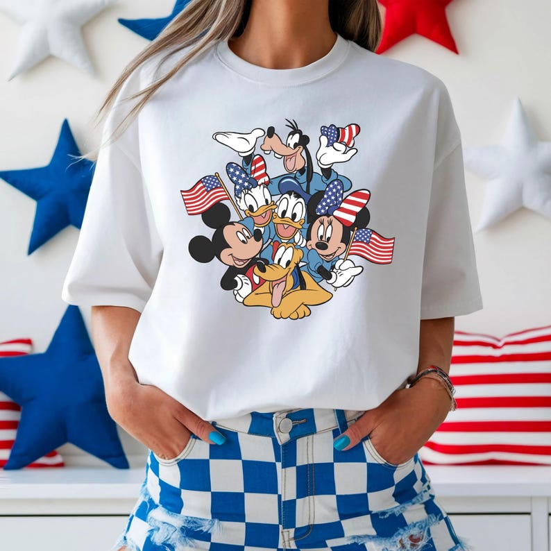 May include: White t-shirt featuring a graphic of Mickey Mouse, Minnie Mouse, Donald Duck, Daisy Duck, Goofy, and Pluto holding American flags. The characters are in patriotic attire. The shirt is paired with blue and white checkered shorts.