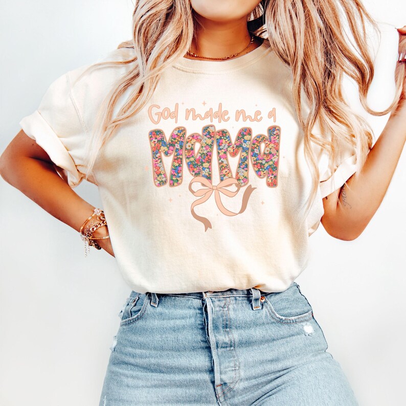 Pu&ograve; includere: T-shirt color crema con la scritta "God made me a Mama" in un design floreale. La parola "Mama" &egrave; riempita di fiori colorati, e sotto c'&egrave; un nastro marrone chiaro. La maglietta &egrave; abbinata a jeans blu.