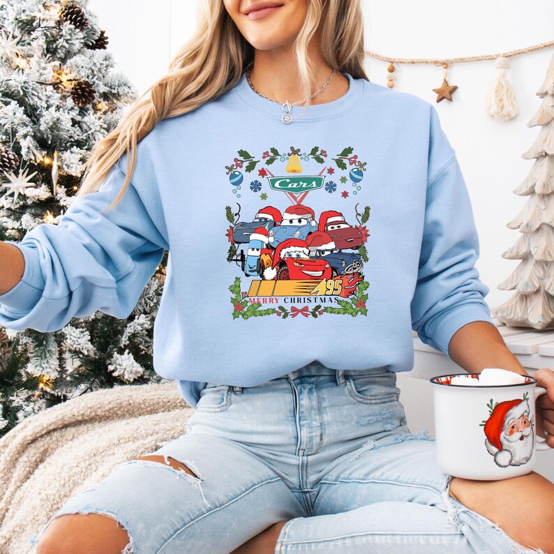 May include: Light blue sweatshirt featuring a graphic of Cars characters wearing Santa hats, with the text "Cars" and "Merry Christmas 95". The design includes festive holly and ornaments. A cozy holiday-themed sweatshirt.