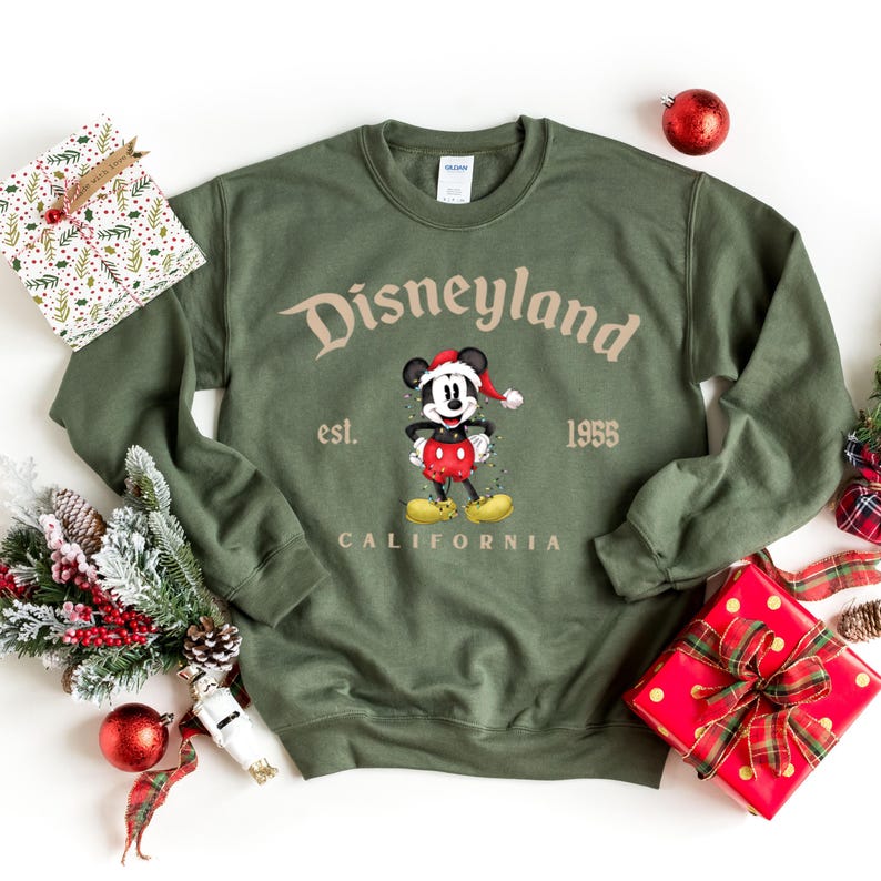 May include: Green sweatshirt with a vintage Disneyland logo featuring Mickey Mouse wearing a Santa hat. The text reads "Disneyland est. 1955 California".
