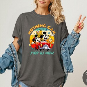 May include: Dark gray t-shirt featuring a colorful graphic of Mickey and Minnie Mouse in a red car, with the text "NOTHING CAN STOP US NOW!". The design is set against a sunset backdrop with butterflies. The shirt is a Comfort Colors Pepper.