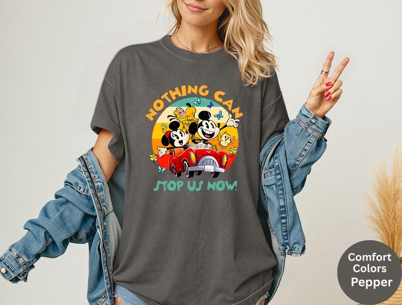 May include: Dark gray t-shirt featuring a colorful graphic of Mickey and Minnie Mouse in a red car, with the text "NOTHING CAN STOP US NOW!". The design is set against a sunset backdrop with butterflies. The shirt is a Comfort Colors Pepper.