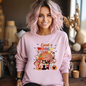 May include: A light pink sweatshirt with the text "Epcot World Tour" and a graphic featuring Disney characters in Halloween costumes. The design includes pumpkins, bats, and the Epcot ball.