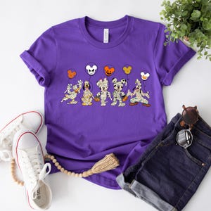Peut inclure: T-shirt violet avec un motif de Mickey Mouse, Minnie Mouse, Donald Duck, Dingo et Pluto habill&eacute;s en momies tenant des friandises d'Halloween. Les personnages tiennent des ballons avec des oreilles de Mickey Mouse et des motifs sur le th&egrave;me d'Halloween.
