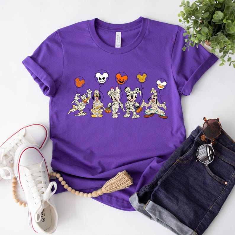 Peut inclure: T-shirt violet avec un motif de Mickey Mouse, Minnie Mouse, Donald Duck, Dingo et Pluto habill&eacute;s en momies tenant des friandises d'Halloween. Les personnages tiennent des ballons avec des oreilles de Mickey Mouse et des motifs sur le th&egrave;me d'Halloween.