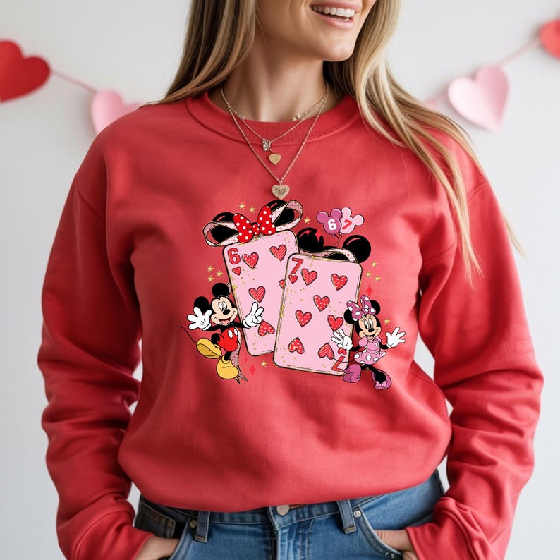 May include: Red sweatshirt featuring a graphic of Mickey and Minnie Mouse with playing cards. The cards are pink with red hearts and the numbers 6 and 7. Mickey Mouse is wearing red shorts and Minnie Mouse is wearing a pink dress.