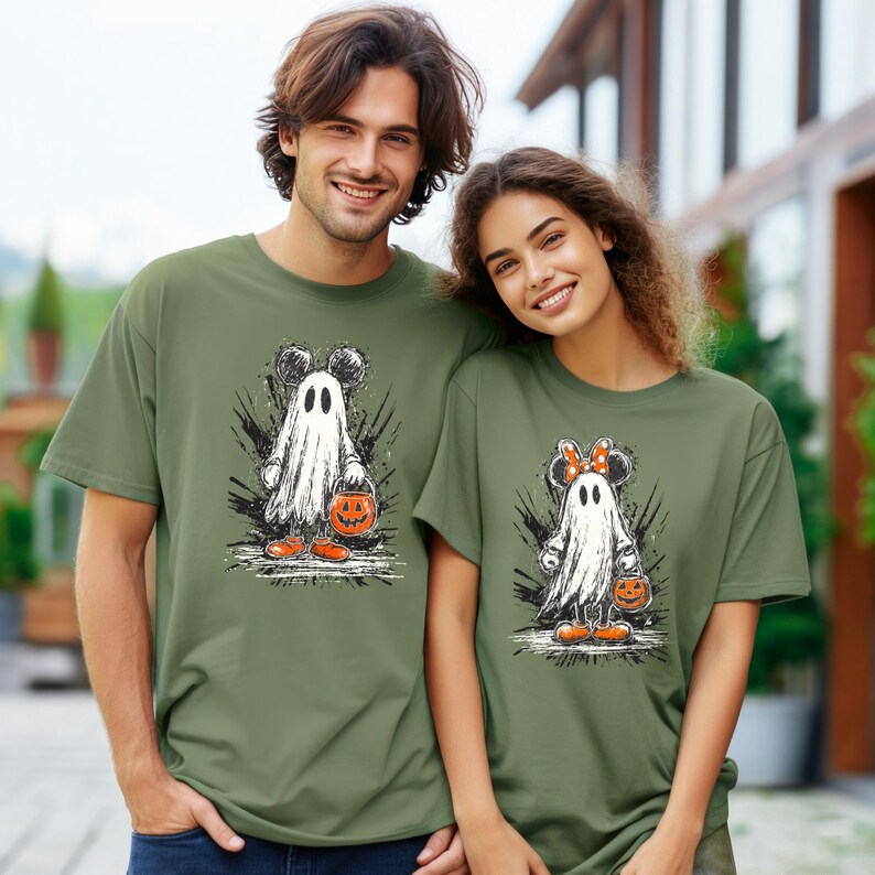 Peut inclure: Deux t-shirts vert olive avec un motif de fant&ocirc;me de dessin anim&eacute;. Le fant&ocirc;me porte des oreilles de Mickey Mouse et un seau de citrouille. Le fant&ocirc;me est blanc avec des accents noirs. Port&eacute;s par un homme et une femme.