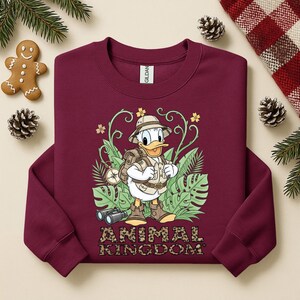 May include: Burgundy sweatshirt featuring a graphic of Donald Duck in safari attire, surrounded by tropical foliage. The text "ANIMAL KINGDOM" is printed in a leopard print font. Includes a gingerbread man and pinecones.