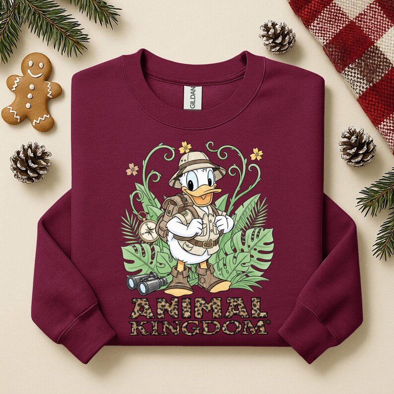 May include: Burgundy sweatshirt featuring a graphic of Donald Duck in safari attire, surrounded by tropical foliage. The text "ANIMAL KINGDOM" is printed in a leopard print font. Includes a gingerbread man and pinecones.