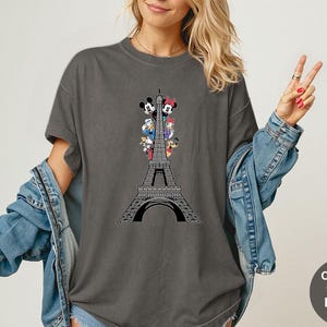 May include: Dark grey t-shirt featuring a black Eiffel Tower design with cartoon characters. The shirt has a crew neck and short sleeves. The text "Comfort Colors Pepper" is on the bottom right.