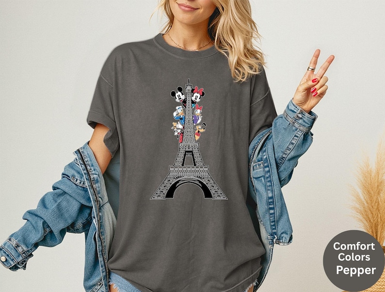 May include: Dark grey t-shirt featuring a black Eiffel Tower design with cartoon characters. The shirt has a crew neck and short sleeves. The text "Comfort Colors Pepper" is on the bottom right.