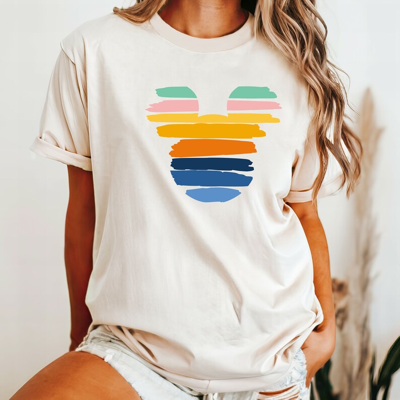 May include: A cream colored t-shirt with a colorful Mickey Mouse silhouette design. The silhouette is made up of horizontal stripes in shades of green, pink, orange, yellow, blue, and light blue.