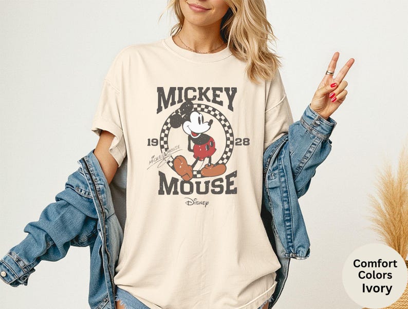 May include: Ivory-colored t-shirt featuring a vintage Mickey Mouse graphic. The design includes the text "Mickey Mouse" and the year "1928" within a circular checkered frame. The shirt has short sleeves and a relaxed fit.