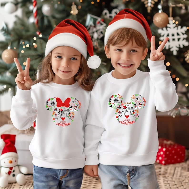 Pu&ograve; includere: Due bambini indossano felpe bianche con un design a forma di Topolino riempito di grafica natalizia e la scritta "MERRY CHRISTMAS". Indossano anche cappelli di Babbo Natale rossi e bianchi. I bambini sorridono e fanno il segno della vittoria.