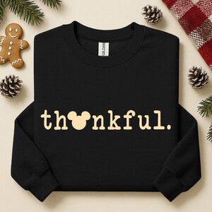 Puede incluir: Sudadera negra con la palabra "thankful" en color beige claro. La "o" de la palabra se reemplaza con una cabeza de Mickey Mouse. La sudadera est&aacute; rodeada de pi&ntilde;as, un hombre de jengibre y vegetaci&oacute;n.