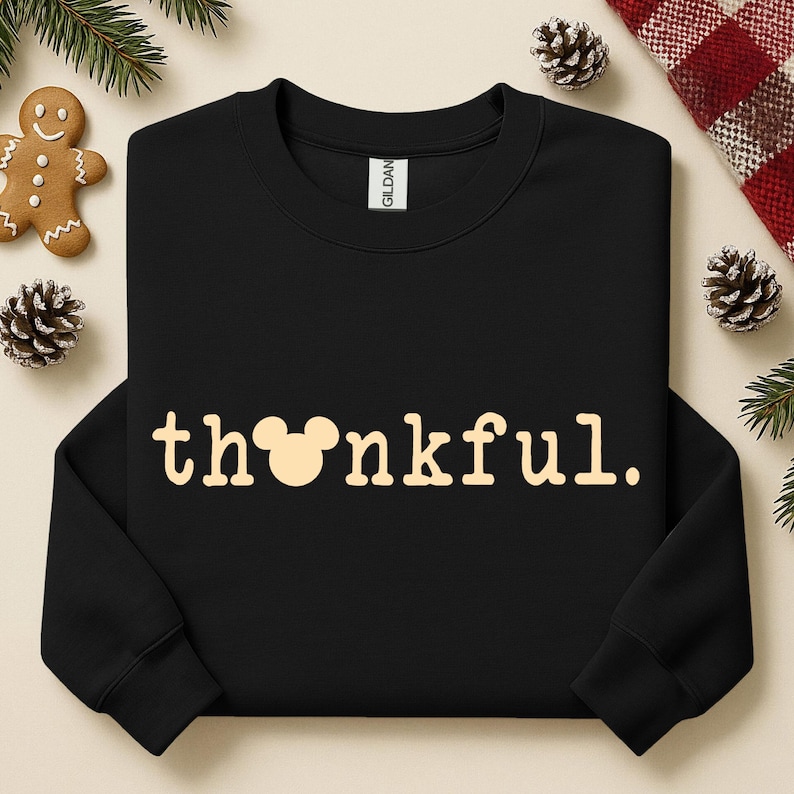 Puede incluir: Sudadera negra con la palabra "thankful" en color beige claro. La "o" de la palabra se reemplaza con una cabeza de Mickey Mouse. La sudadera est&aacute; rodeada de pi&ntilde;as, un hombre de jengibre y vegetaci&oacute;n.