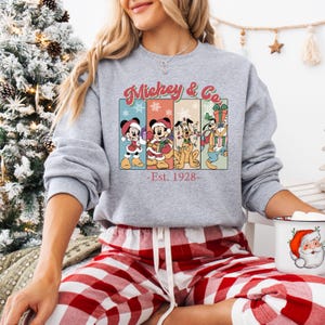 May include: A gray sweatshirt with a graphic featuring Mickey Mouse, Minnie Mouse, Pluto, and Goofy in Christmas attire. The text "Mickey & Co. - Est. 1928-" is displayed above the characters. The person is wearing red and white plaid pajama pants.
