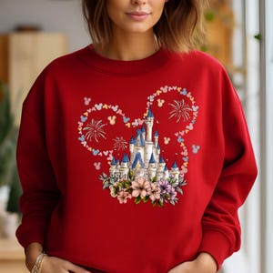 May include: Red sweatshirt featuring a castle design with floral accents and a Mickey Mouse ear outline. The design includes fireworks and Mickey Mouse silhouettes in pastel colors. The sweatshirt is a crew neck style.