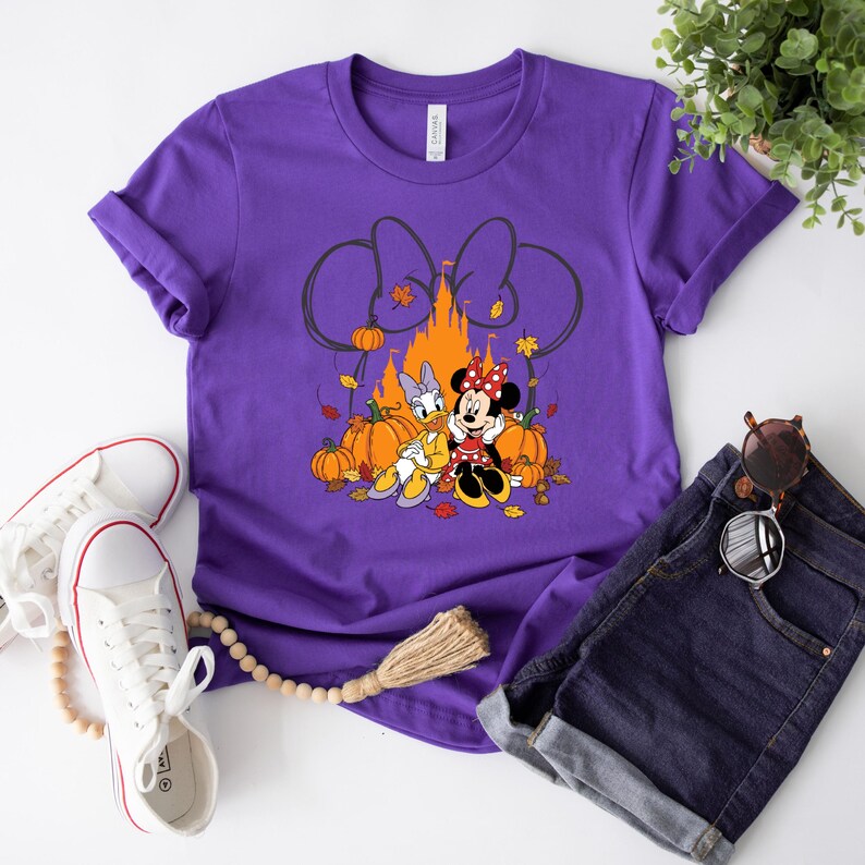 May include: Purple t-shirt featuring a graphic of Minnie Mouse and Daisy Duck with pumpkins, autumn leaves, and a castle. The design includes a large bow and the word "Canvas" on the tag. The shirt is paired with denim shorts and white sneakers.