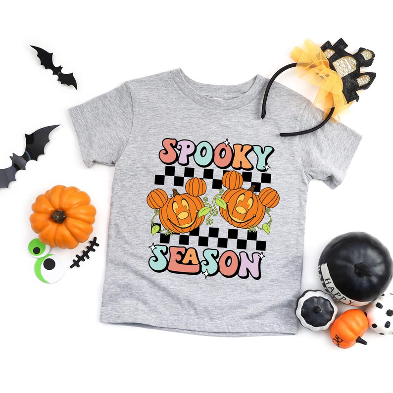 May include: A light gray t-shirt with the words "Spooky Season" in colorful, retro-style lettering. The shirt features two orange pumpkin faces with mouse ears on a black and white checkered background. Halloween-themed.