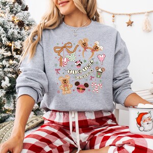 May include: A grey sweatshirt with a Christmas-themed design. The graphic includes gingerbread Mickey Mouse heads, candy canes, and the text "Tis the Season." The design incorporates pink, brown, and green colours, ideal for the festive period.