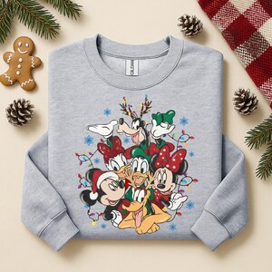 May include: A light gray sweatshirt featuring a festive graphic of Disney characters, including Mickey and Minnie Mouse, Donald and Daisy Duck, Goofy, and Pluto, all adorned with holiday decorations and string lights.