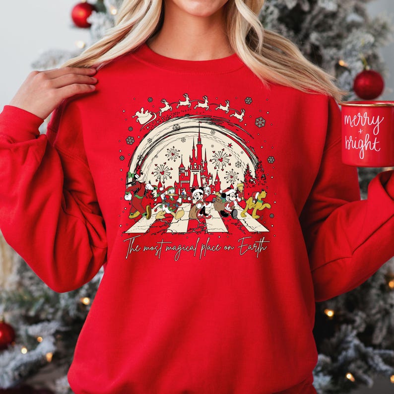 May include: Red Christmas sweatshirt with a white graphic of Mickey Mouse and friends walking across a crosswalk. The graphic includes the words 'The most magical place on Earth' and a silhouette of Cinderella's castle with fireworks. The sweatshirt has a crew neck and long sleeves.