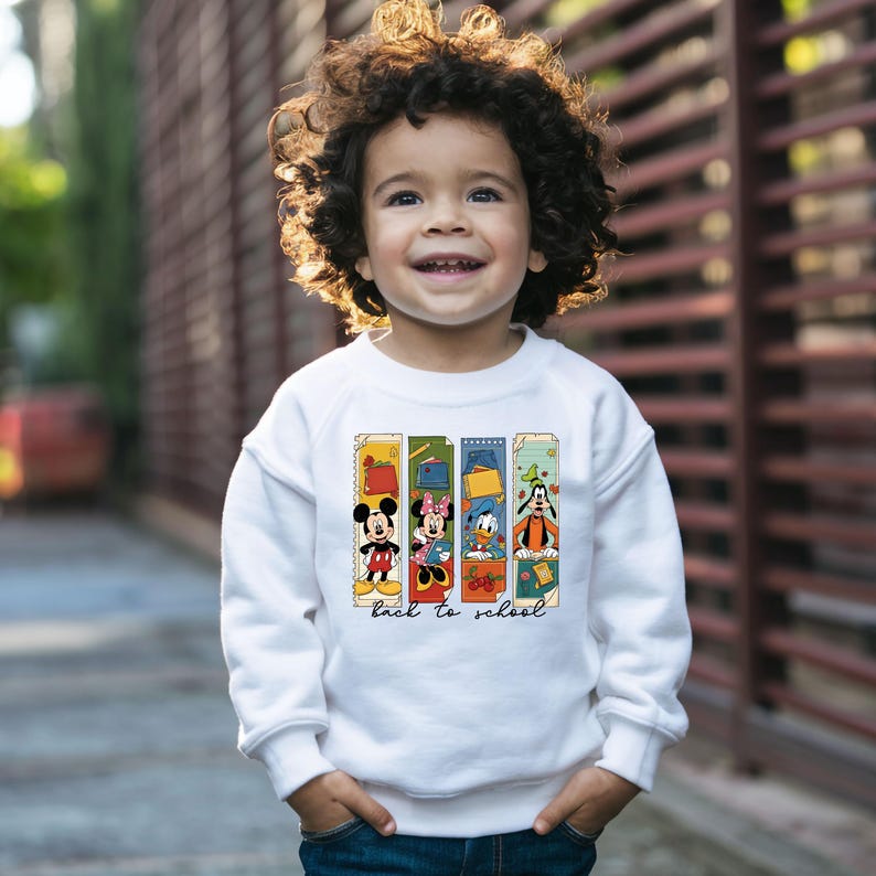 May include: A white sweatshirt featuring Mickey Mouse, Minnie Mouse, Donald Duck, and Goofy in a back-to-school design. The graphic includes the text "back to school" in a script font. The child is smiling.