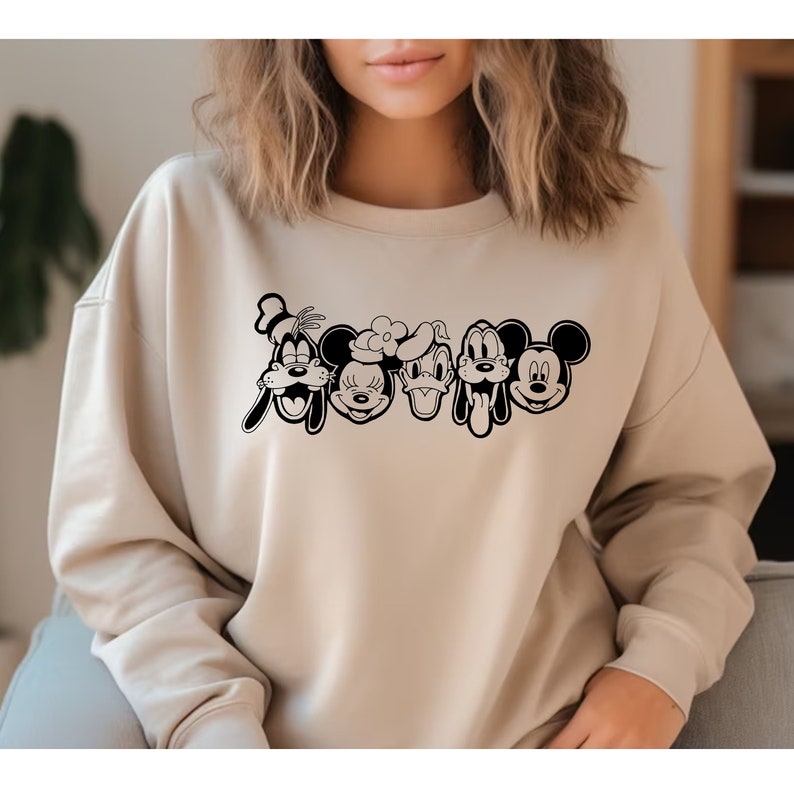 K&ouml;nnte beinhalten: Ein beiger Pullover mit einem schwarzen Grafikdruck von Mickey Mouse, Minnie Mouse, Donald Duck, Goofy und Pluto. Die Figuren sind in einer Reihe angeordnet, nach vorne gerichtet, mit offenem Mund.