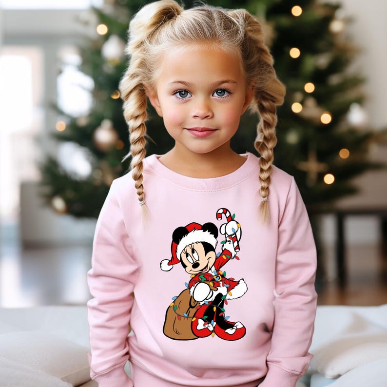 May include: A light pink sweatshirt featuring a Christmas-themed Mickey Mouse graphic. Mickey is wearing a Santa hat and suit, holding a candy cane and a sack of presents, and is wrapped in Christmas lights. The sweatshirt is worn by a young girl.