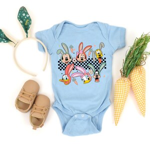 May include: Light blue baby onesie with a graphic of cartoon characters dressed as bunnies. The characters include Mickey Mouse, Minnie Mouse, Donald Duck, Goofy, and Pluto. The onesie has a checkered pattern and is surrounded by carrots and Easter grass.