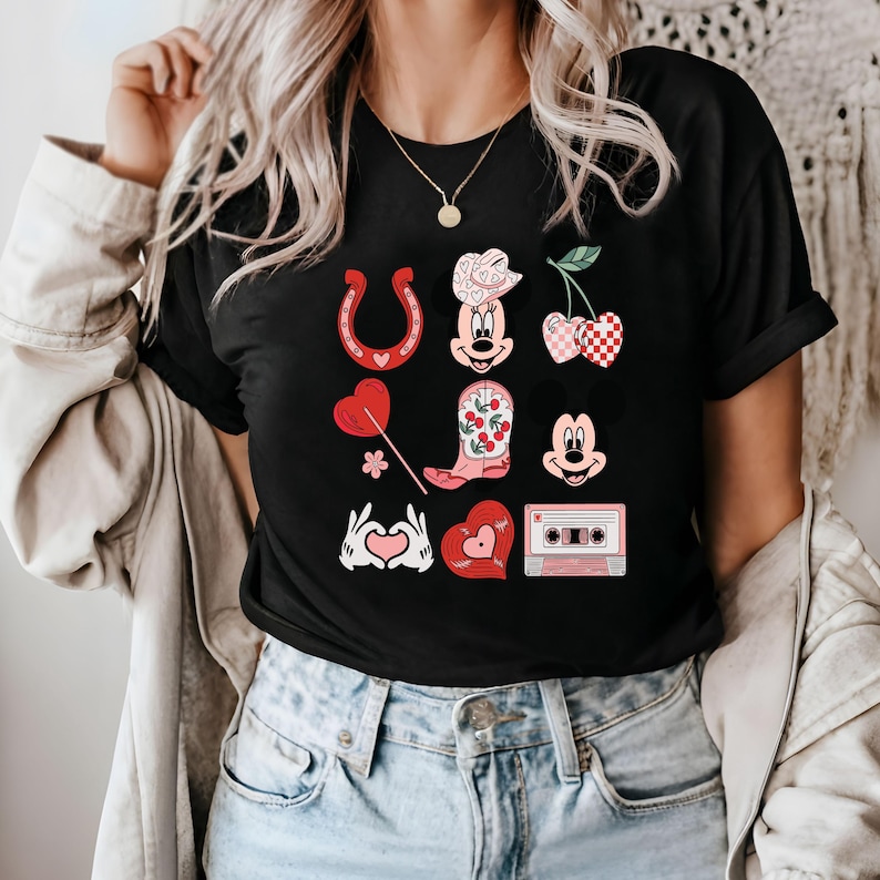 May include: Black t-shirt featuring a graphic print of a cartoon character wearing a cowboy hat and boots, a horseshoe, a cherry, a heart, a flower, and a cassette tape.