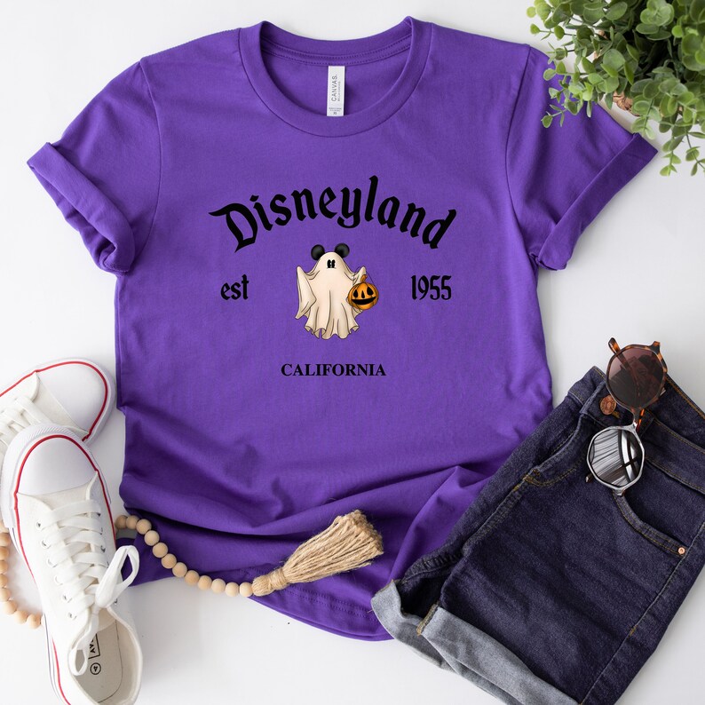 Puede incluir: Camiseta morada con un gr&aacute;fico negro de un fantasma sosteniendo una calabaza. El texto "Disneyland est 1955 CALIFORNIA" est&aacute; impreso en la camiseta.