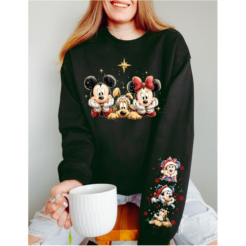 May include: Black sweatshirt featuring Mickey Mouse, Pluto, and Minnie Mouse in festive attire. The design includes a star and additional character graphics on the sleeve. A white mug is held in the person's hand.