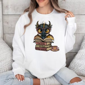 May include: White sweatshirt with a black dragon wearing glasses and reading a book. The dragon is sitting on a stack of books with the text "read books be kind stay weird". There is a cup of coffee next to the books.
