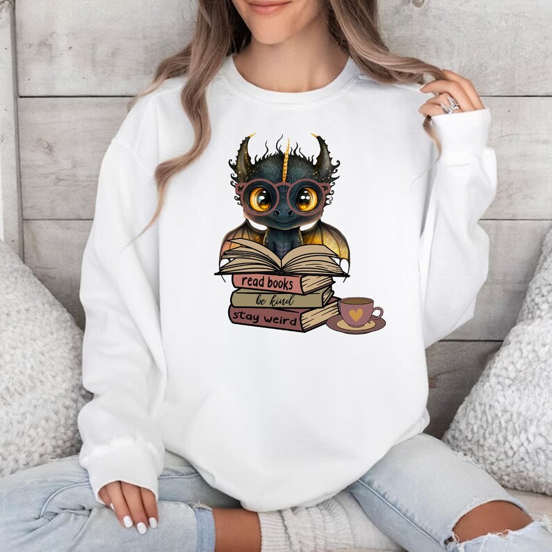May include: White sweatshirt with a black dragon wearing glasses and reading a book. The dragon is sitting on a stack of books with the text "read books be kind stay weird". There is a cup of coffee next to the books.