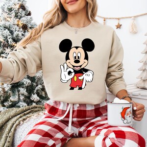 May include: A beige sweatshirt with a Mickey Mouse graphic, showing him making an "OK" hand gesture. Mickey is wearing his iconic red shorts, white gloves, and has black ears. The sweatshirt is worn with red and white check pajama bottoms.