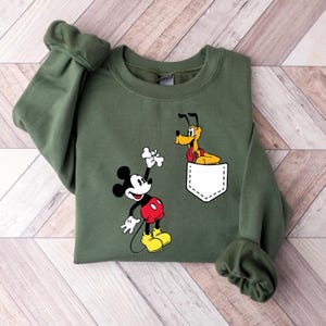 Puede incluir: Una sudadera verde con un bolsillo blanco con Mickey Mouse y Pluto. Mickey Mouse sostiene un hueso y Pluto asoma por el bolsillo.