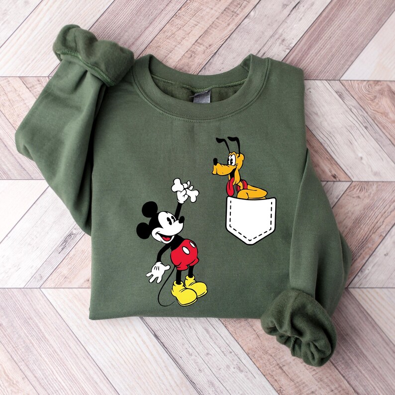 Puede incluir: Una sudadera verde con un bolsillo blanco con Mickey Mouse y Pluto. Mickey Mouse sostiene un hueso y Pluto asoma por el bolsillo.