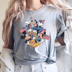 May include: Gray t-shirt featuring a graphic of Mickey Mouse, Minnie Mouse, Donald Duck, Daisy Duck, Goofy, and Pluto, all holding American flags. The characters are in patriotic attire. The shirt is a crew neck style.