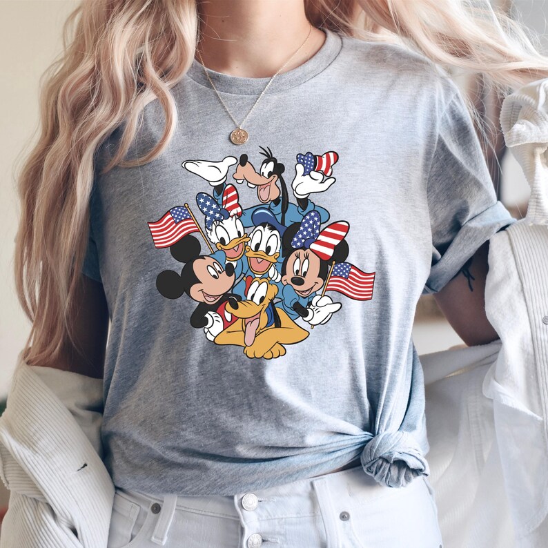 May include: Gray t-shirt featuring a graphic of Mickey Mouse, Minnie Mouse, Donald Duck, Daisy Duck, Goofy, and Pluto, all holding American flags. The characters are in patriotic attire. The shirt is a crew neck style.