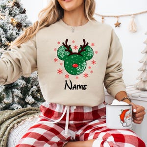Może przedstawiać: Beżowy świąteczny sweter z zielonym wzorem głowy renifera Mickey Mouse i śnieżynkami. Sweter ma napis "NAME" nadrukowany na dole.