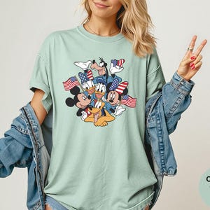 May include: A light green t-shirt featuring a graphic of Mickey Mouse, Minnie Mouse, Donald Duck, Goofy, and Pluto, each holding an American flag. The shirt is a Comfort Colors Bay style.