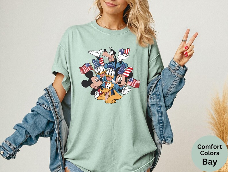 May include: A light green t-shirt featuring a graphic of Mickey Mouse, Minnie Mouse, Donald Duck, Goofy, and Pluto, each holding an American flag. The shirt is a Comfort Colors Bay style.