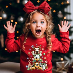 Peut inclure: Une jeune fille blonde aux yeux bleus porte un pull de No&euml;l rouge et un n&oelig;ud rouge. Le pull repr&eacute;sente Mickey Mouse, Donald Duck et Pluto avec des d&eacute;corations de vacances. Un sapin de No&euml;l est en arri&egrave;re-plan.