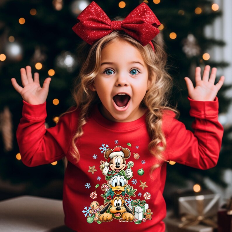 Peut inclure: Une jeune fille blonde aux yeux bleus porte un pull de No&euml;l rouge et un n&oelig;ud rouge. Le pull repr&eacute;sente Mickey Mouse, Donald Duck et Pluto avec des d&eacute;corations de vacances. Un sapin de No&euml;l est en arri&egrave;re-plan.