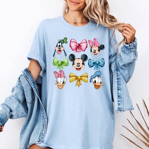May include: Light blue t-shirt with a graphic print of various Disney characters wearing colorful bows. The characters include Mickey Mouse, Minnie Mouse, Donald Duck, Goofy, and Daisy Duck.