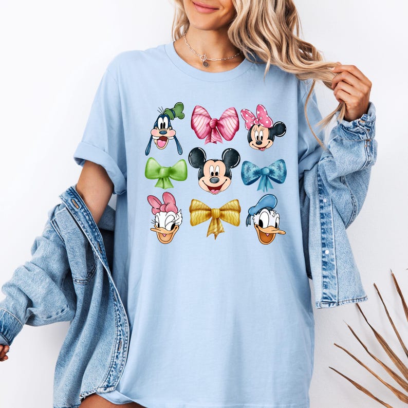 May include: Light blue t-shirt with a graphic print of various Disney characters wearing colorful bows. The characters include Mickey Mouse, Minnie Mouse, Donald Duck, Goofy, and Daisy Duck.